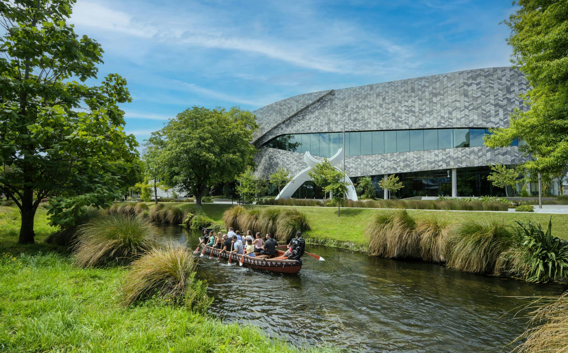 A group paddles a canoe along a lush river, bordered by greenery and tall grass. In the background, a modern building with a distinctive patterned facade is visible under a blue sky.